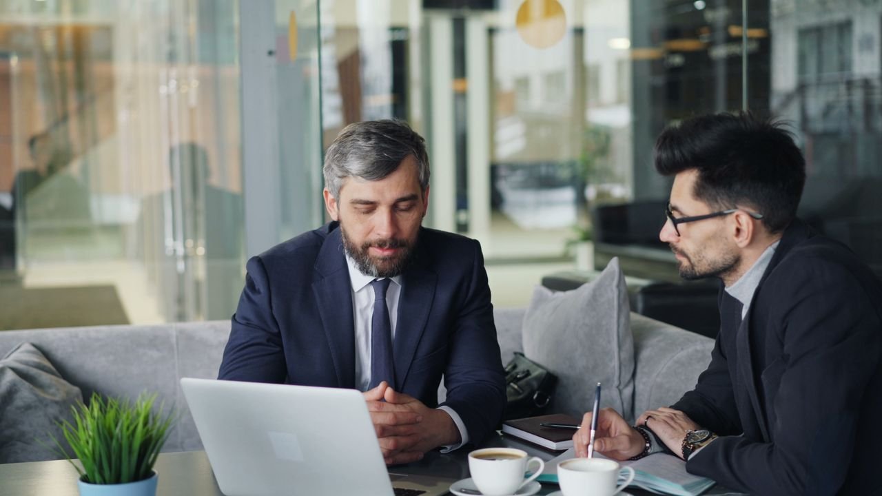 Mastering the First Impression: Your intriguing post title goes here Two businessmen in suits discussing work at a cafe with laptops and coffee.