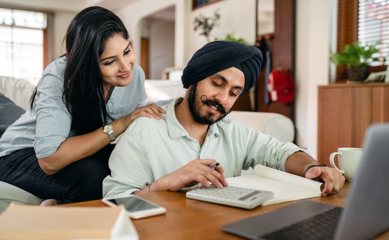 Portfolio Concentrated ethnic male accountant using calculator with gadgets and books and notebook for remote work while ethnic wife touching shoulder of husband watching for numbers behind back sitting on sofa in living room
