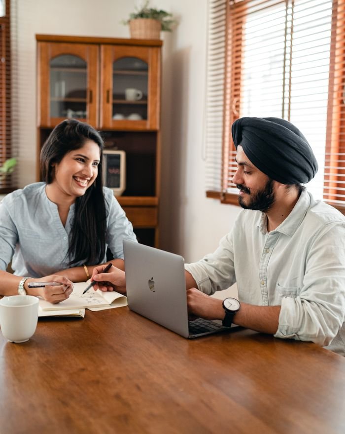 Our Services Smiling young Indian man in shirt and turban using laptop at wooden table and helping cheerful female friend while working remotely at home