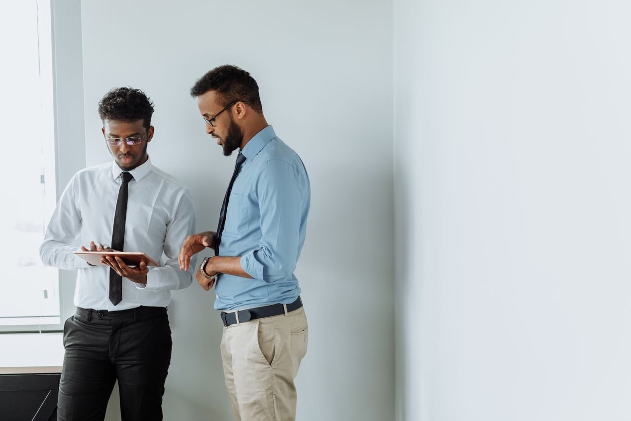 Portfolio Two businessmen discussing work with a tablet against a white wall in a modern office setting.